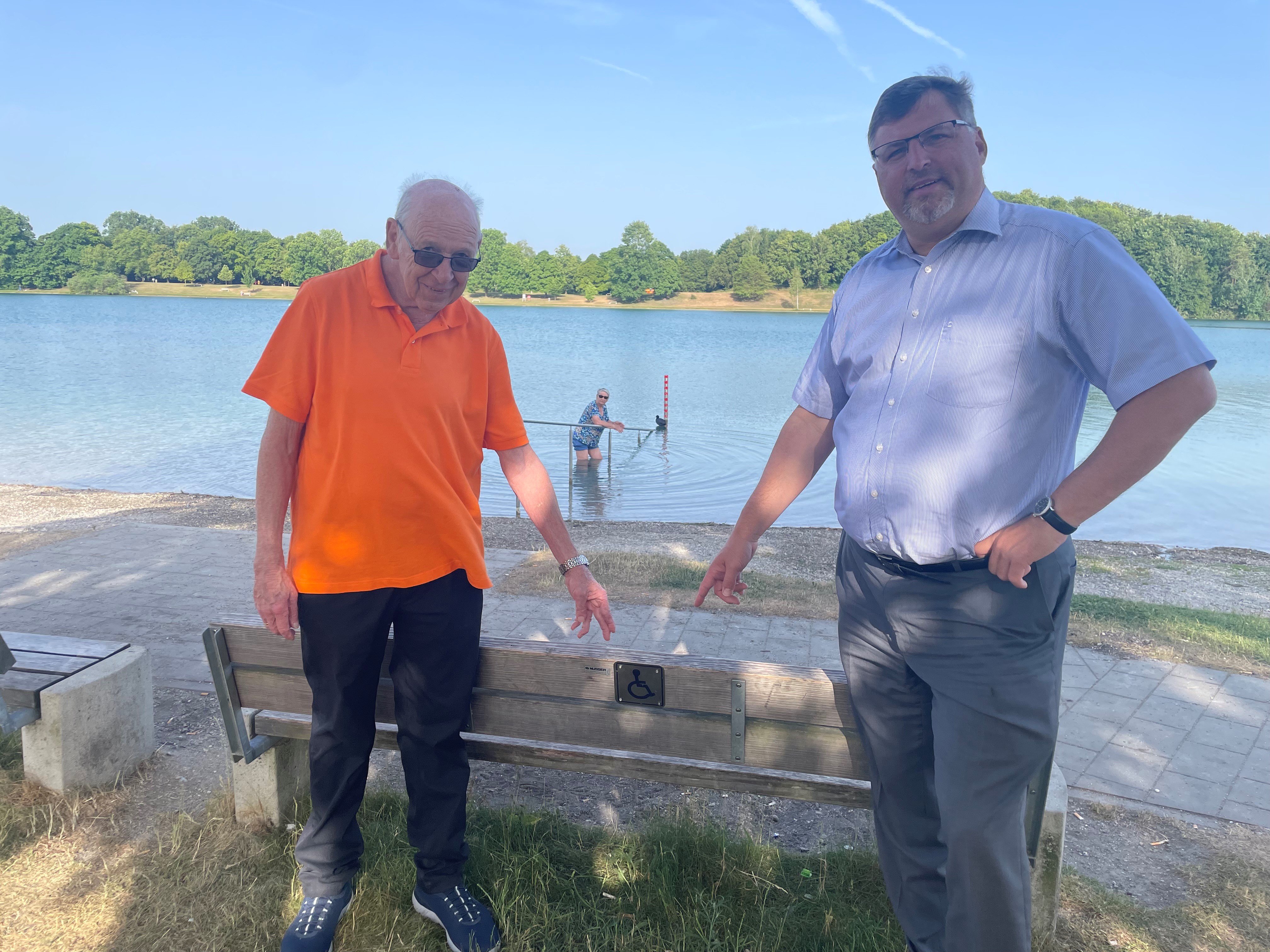 Landrat Stefan Löwl und Hartmut Baumgärtner, Kreisbeauftragter für Menschen mit Behinderung im Landkreis Dachau an einer der fünf Bänke am Karlsfelder See, die nun per Schild Menschen mit Behinderung Vorrang gewährt.