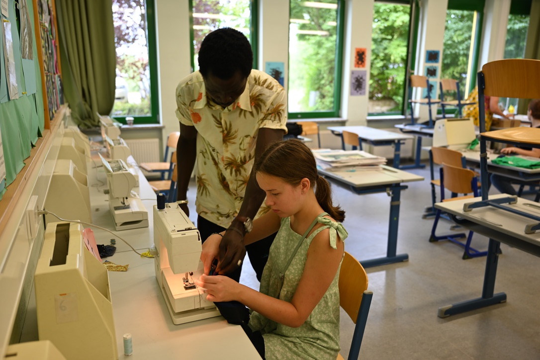 Ein Schneider aus Afrika zeigt einer Schülerin an der Nähmaschine wie sie aus gebrauchtem Stoff etwas Neues entstehen lassen kann.