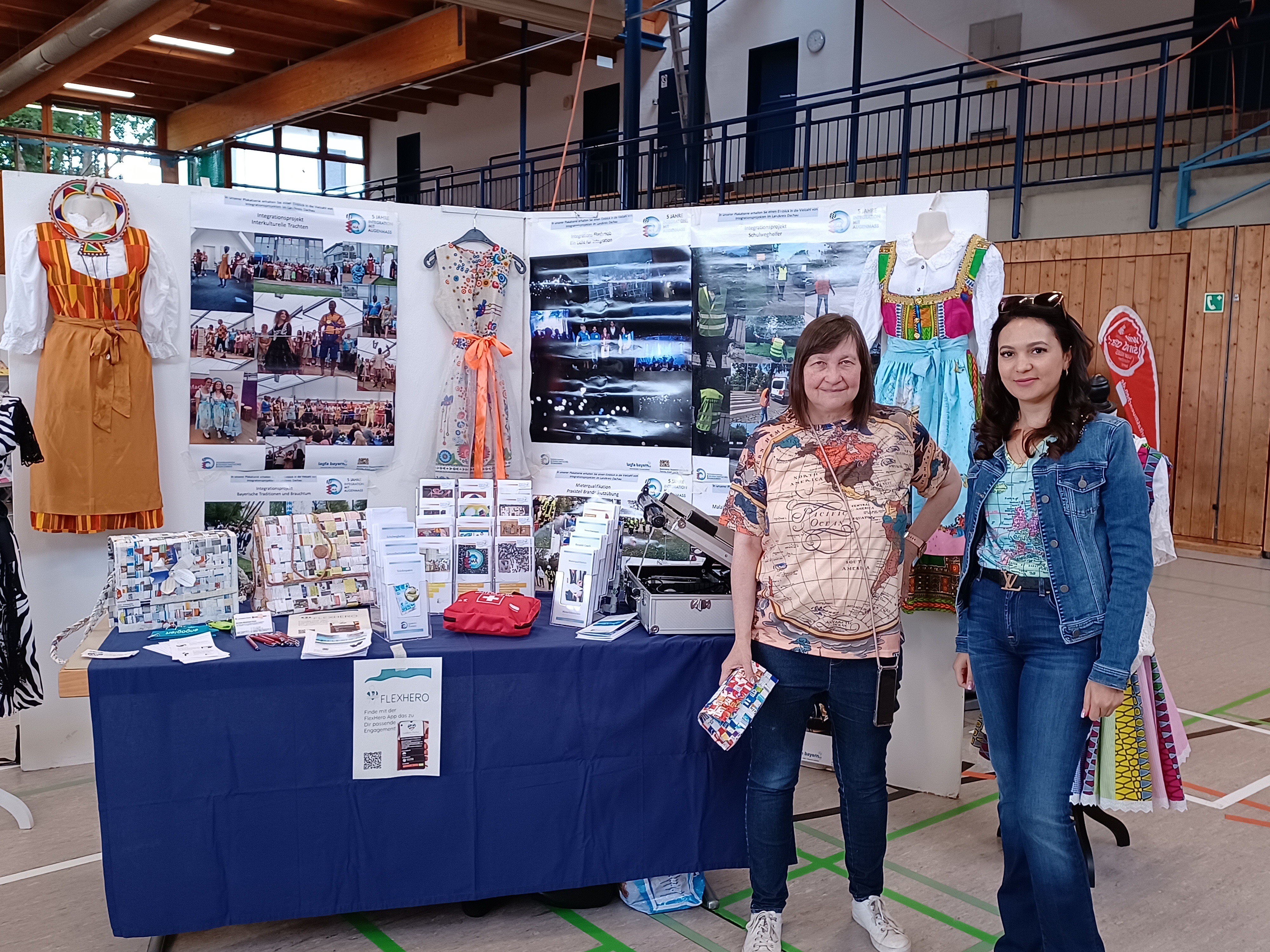 Frau Wintermayr-Greck und Frau Keller von IMA Team vor dem bunten Stand auf der Gesundheitsmesse. Zusehen sind unter anderem selbstgebastelte Papiertaschen und bunte Kleider im afrikanisch-bayrischen Design.