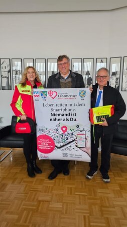 Bild vergr&ouml;&szlig;ern: Drei Personen stehen in einem Raum und halten ein großes Plakat der Initiative „Region der Lebensretter“. Auf dem Plakat steht: „Leben retten mit dem Smartphone. Niemand ist näher als Du.“ Links hält eine Frau eine rote Notfalltasche, rechts ein Mann eine gelb-rote Notfallausrüstung. Die Aktion wirbt dafür, eine App herunterzuladen und sich als Ersthelferin oder Ersthelfer zu registrieren, um bei Notfällen in der Nähe schnell helfen zu können.