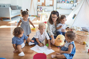 Eine wunderschöne Erzieherin und eine Gruppe von Kleinkindern sitzen im Kindergarten auf dem Boden und zeichnen mit Papier und Bleistift, umgeben von zahlreichen Spielsachen.
