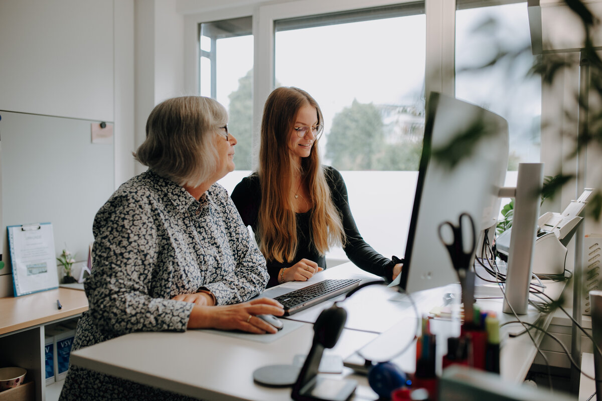 Zwei Kolleginnen stehen zusammen an einem Schreibtisch und arbeiten gemeinsam an einem Bildschirm
