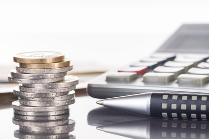 Gestapelte Euromünzen neben Taschenrechner und Stiften auf einem Schreibtisch.