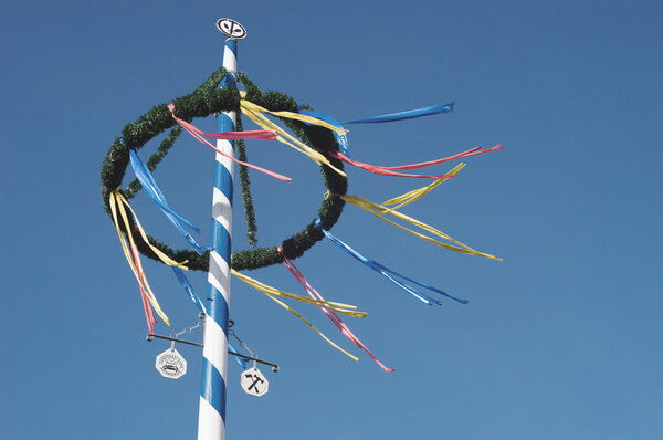 Oberer Teil eines Maibaums mit farbenfrohen Bändern vor einem strahlend blauen Himmel.“